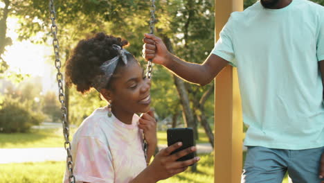 Amigos-Afroamericanos-Chateando,-Usando-Un-Teléfono-Inteligente-Y-Columpiándose-En-El-Parque
